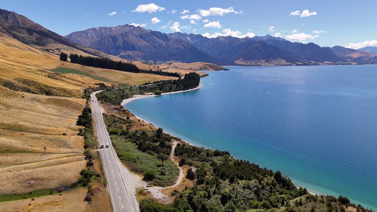 4k drone video of cars and camper vans driving along the road next to Lake Hawea in Wanaka, New Zealand. The vast blue lake is surrounded by fields, hills and mountains