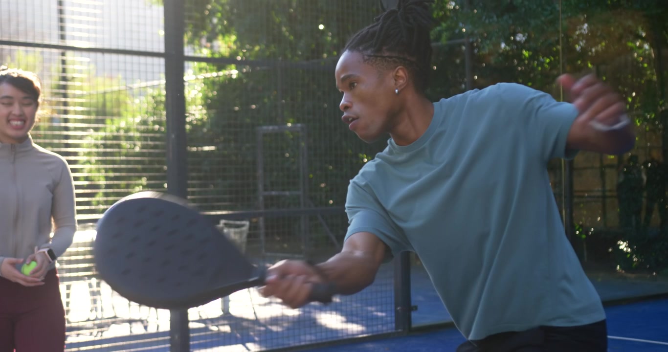 African American man and woman training on glass court, woman dropping ball man swinging paddle