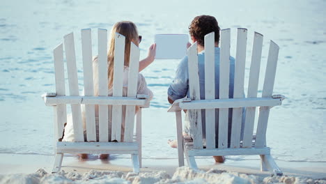 Young-couple-in-chaise-longues-holding-pad-and-looking-at-it