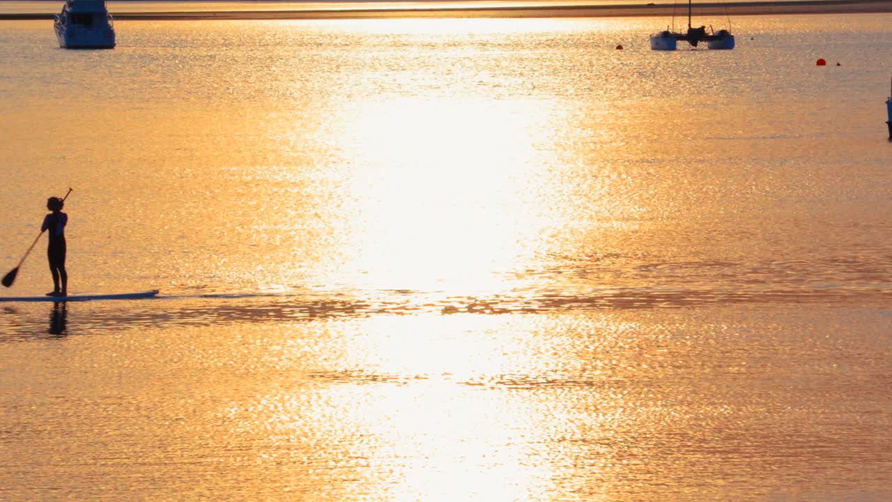Female stand up paddle boarder passing through frame with golden sunlight on water