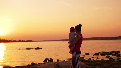 woman holding a baby in her arms stands at the river bank at sunset. happy family time spent in the nature. pink sky backdrop.