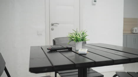 modern dining table with black wooden surface featuring a small potted plant, remote control, and smartphone. a white door in the background leads to a well lit space with minimalistic design