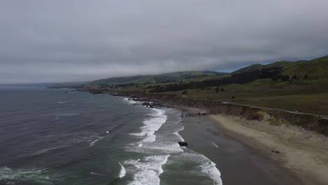 cars drive by on the coastal highway as waves crash onto the beach in a coastal town in northern california. the ocean is contrasted by quiet, green hills in the background. 4k cinematic drone footage