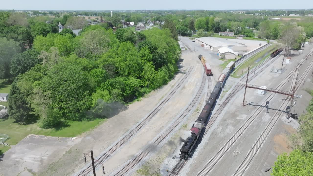 A vintage freight steam train chugs along the tracks surrounded by vibrant greenery and residential areas