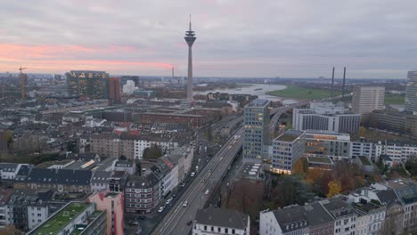 Vista-Aérea-De-Drones-Sobre-El-Centro-De-Dusseldorf-Hacia-El-Puerto-De-Medios-Y-La-Torre-Del-Rin-Al-Atardecer-Con-Un-Cielo-Colorido