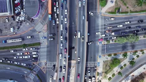 ascenso aéreo sobre el tráfico en movimiento, con vehículos que navegan en múltiples direcciones y estructuras elevadas que aparecen en el horizonte, capturado durante el día en una intersección urbana dinámica