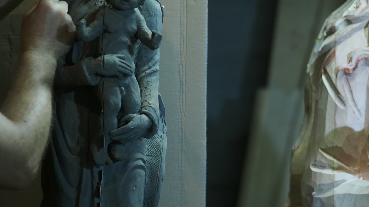 인물의 섬세한 점토 조각상을 손으로 조각하고, 오른쪽에는 부분적으로 형성된 또 다른 조각상이 보이며, 중립적인 배경을 사용하여 창작의 예술적 과정을 보여줘요.