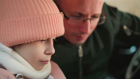 a close-up shot of a young girl in a pink beanie and jacket, sitting quietly in a car, with a man, likely her father, leaning in closely and attentively looking at the phone she is holding