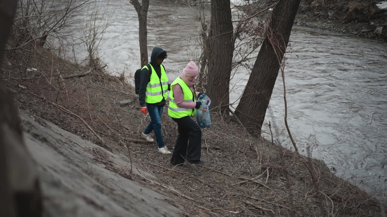 Volunteers in high visibility vests collecting trash along riverbank during environmental cleanup in cold early spring forest with bare trees, overcast sky, and flowing water, promoting community service