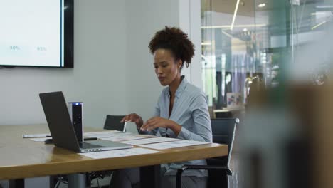 mixed race businesswoman sitting at desk, checking documents and using laptop in office