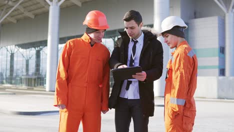 Investor-of-the-project-in-a-black-suit-examining-the-building-object-with-construction-workers-in-orange-uniform-and-helmets.-They-are-cheking-the-drawings-using-a-tablet