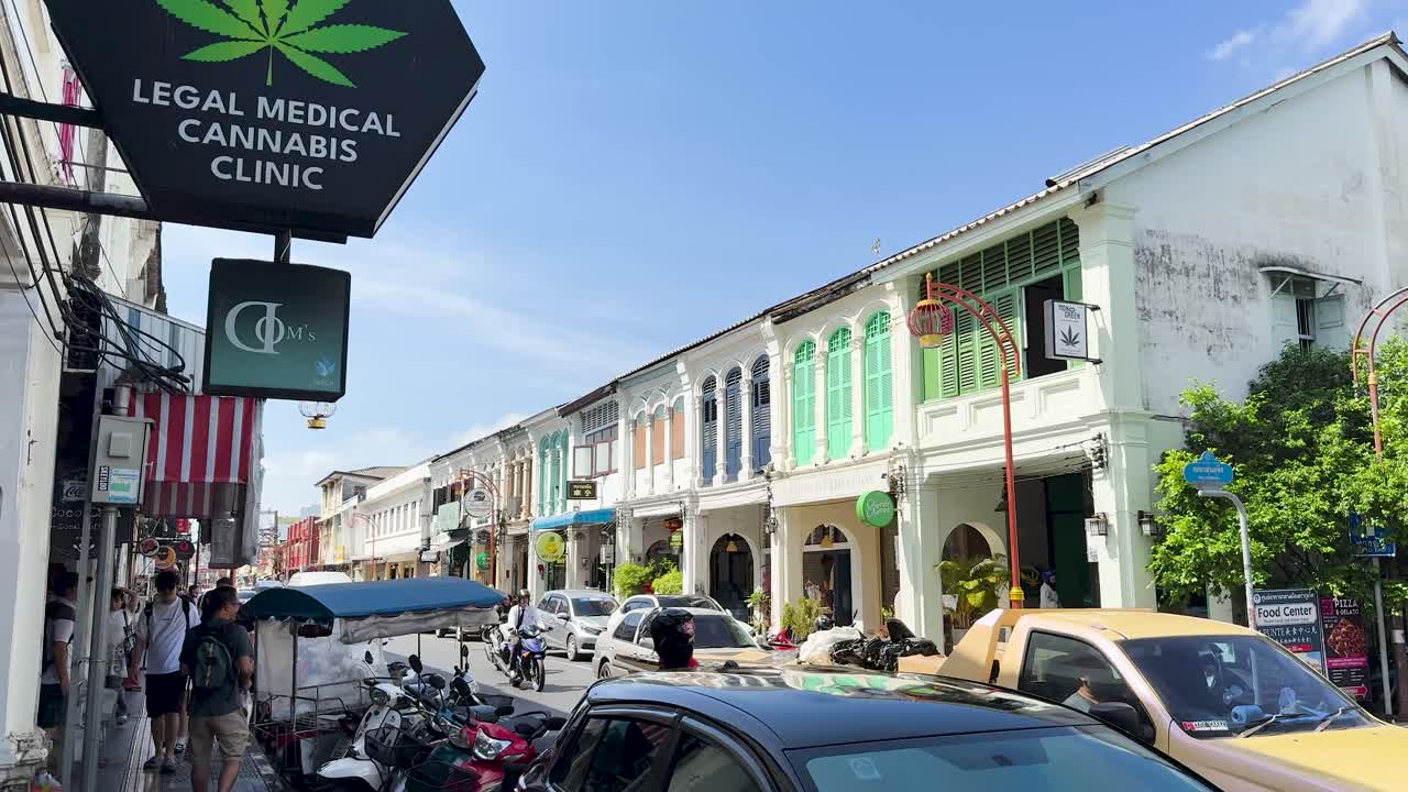 Daytime street view with cannabis clinic sign, colonial buildings, moving vehicles, and pedestrians in Phuket