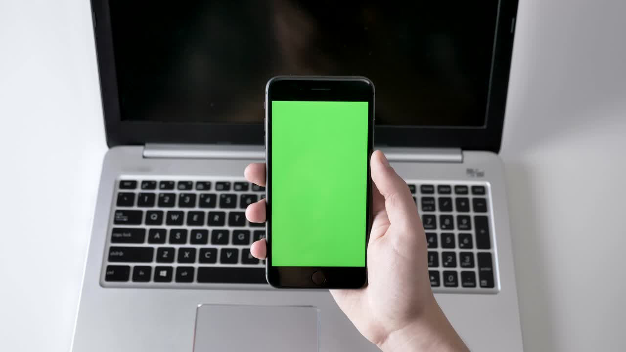 Male hands tap on a larger black smartphone, laptpo in the background. Green screen, chromakey concept, laptop in the background. Top shot 60 fps