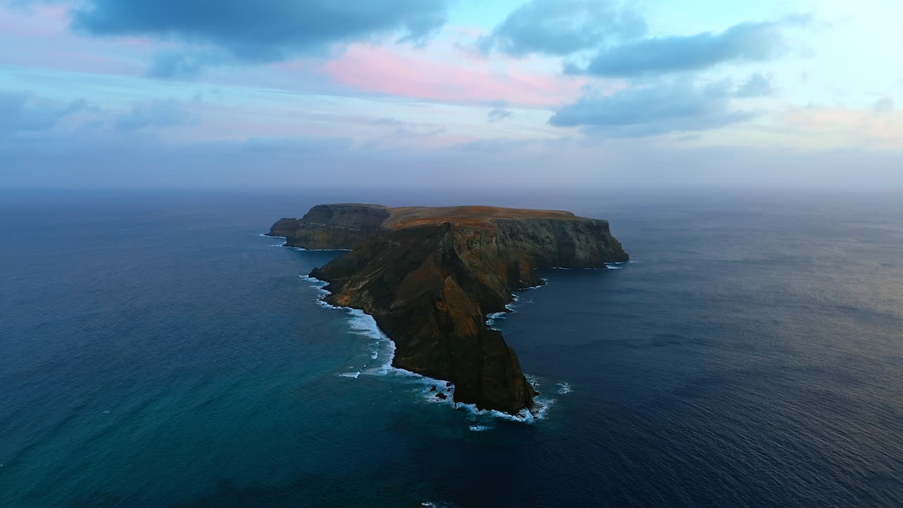 Little rocky island with a plain on the top. Vast Atlantic Ocean surrounding the island. Heavy clouds hang in the sky. Aerial view.
