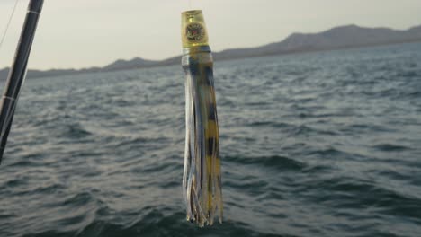close-up of colorful bait lure hanging from a fishing rod with the ocean and mountains in the background.