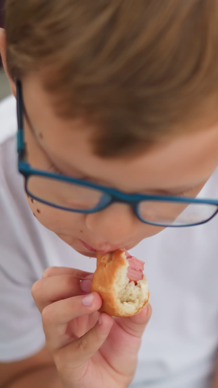 A young boy wearing glasses is eating a sausage roll while looking thoughtfully to his right