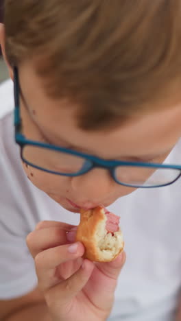 un niño con gafas está comiendo un rollo de salchichas mientras mira pensativo a su derecha