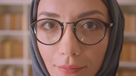 closeup portrait of young muslim female face in glasses and hijab looking at camera smiling cheerfully in library indoors