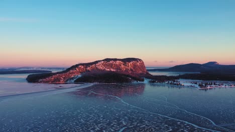 aerial view dropping down towards the ice with a cliff faced mountain lit by a winter sunrise