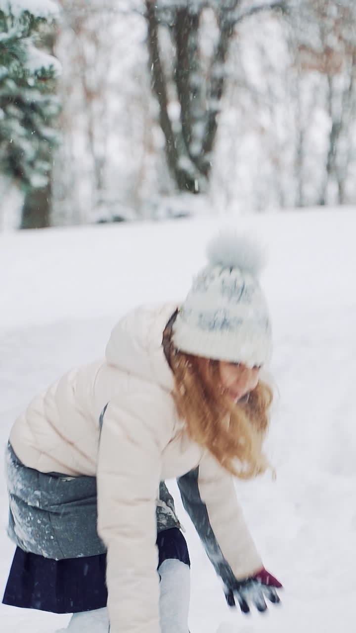 Smiling girl with long hair is throwing snowballs in the street in the yard during a snowfall in the winter. Slow motion Vertical video