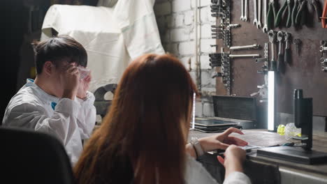 자동차 작업실에 앉아있는 실험실 기술자, 구성 요소를 작업하고, 도구와 장치를 사용하여, 조직된 장비, 형광 조명 및 기계적 도구로 둘러싸여 있습니다.