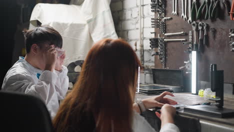 lab technicians seated in automotive workshop working on components, using tools and devices, surrounded by organized equipment, fluorescent lighting, and mechanical instruments