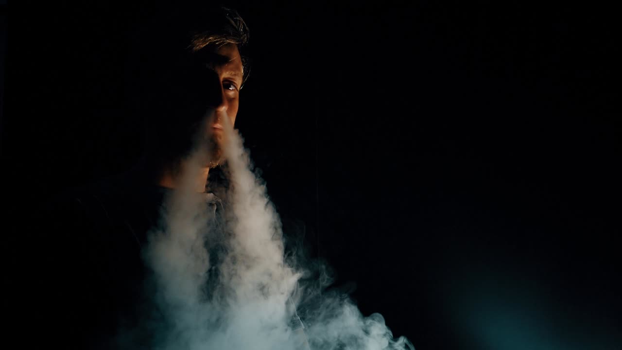 Joven vapeando y exhalando una gran nube de vapor en una habitación oscura