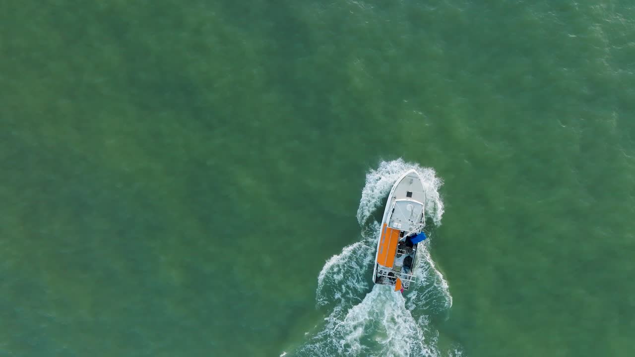 Aerial view of a coastal fishing boat sailing in the calm Baltic sea, fisherman dropping fishing net in the water, sunny day, distant white sand beach, wide birdseye drone shot