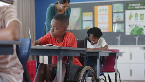 in school, boy in wheelchair writing in notebook with teacher assisting