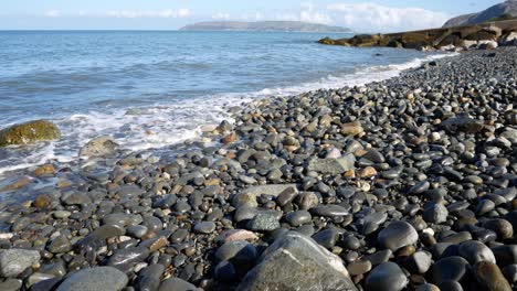 Sunny-blue-ocean-waves-wash-over-stone-pebble-beach-coastline-island-holiday-scene-left-dolly