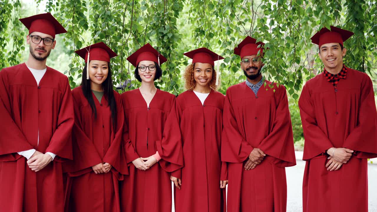 Portrait of multiethnic group of graduating students standing outdoors wearing red gowns and mortar-boards, smiling and looking at camera.