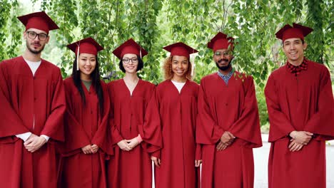 portrait of multiethnic group of graduating students standing outdoors wearing red gowns and mortar-boards, smiling and looking at camera.