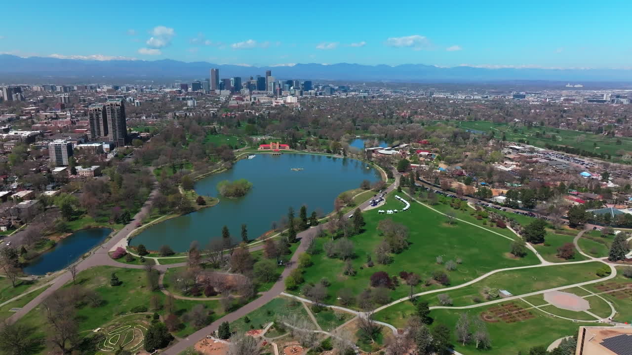City Park Denver Colorado Lake Pavilion vibrant spring summer aerial drone sunny blue sky snow capped Rocky Mountains front range cityscape green lush grass trees blossom pan left motion