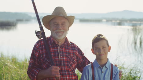 portrait of a gray haired fisherman with his grandson standing at the lake shore, looking at each other and smiling to the camera