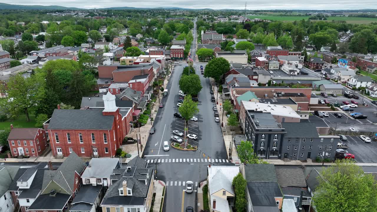 Parking cars in city center of Manheim With waving flag of USA on flagpole. Sunny day with green trees in spring. Historic buildings, houses and church. Descend drone wide shot.