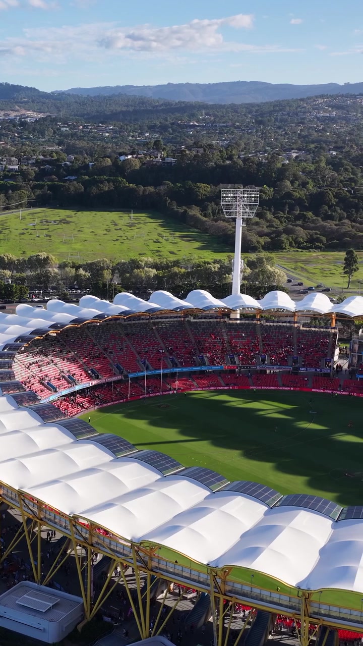 High-angle drone shot pans across a large outdoor stadium under bright daylight and blue skies.
