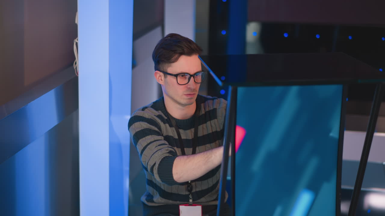 man adjusts digital display screen in studio, focused on setup for video production, wearing striped sweater and ID badge, with blue lights and equipment in background, preparing stage