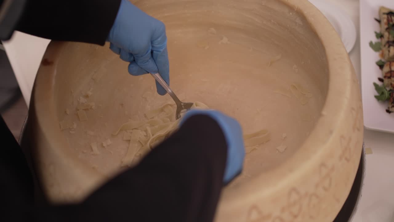 Person scraping cheese from a large cheese wheel with a spoon, wearing blue gloves