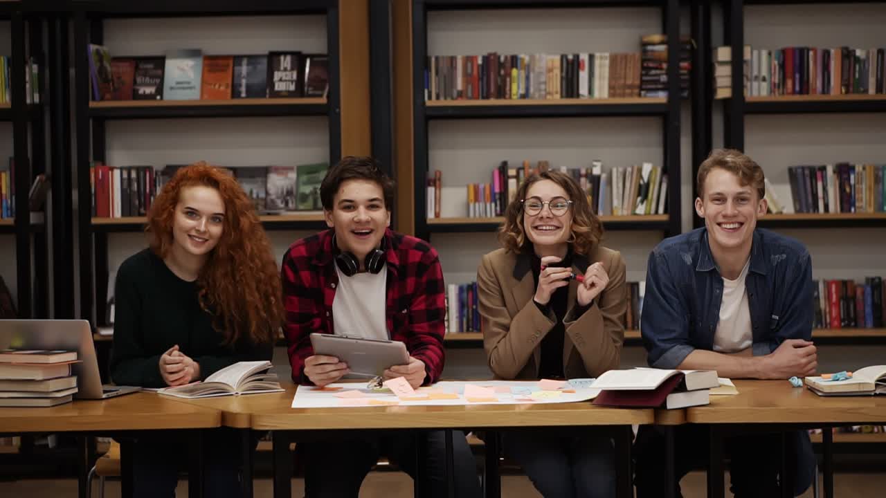Two young male and two female european students sitting at the table with books and laptop in the library and cheerfully smiling. Bookshelves in library with lots of books in background