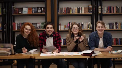 two young male and two female european students sitting at the table with books and laptop in the library and cheerfully smiling. bookshelves in library with lots of books in background