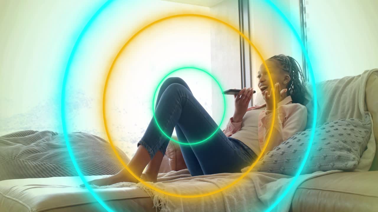Woman reclining on sofa holding device and speaking, tech rings pulsing and shifting hue with voice