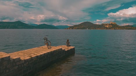 back view of lonely woman with bicycle behind sitting on pier edge on maggiore lake, italy