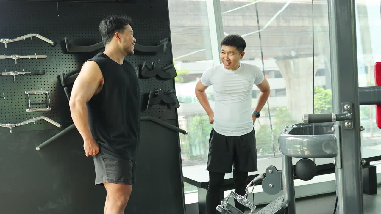 Two men engage in a friendly conversation at a gym, surrounded by equipment. Bright lighting and casual attire create a relaxed atmosphere