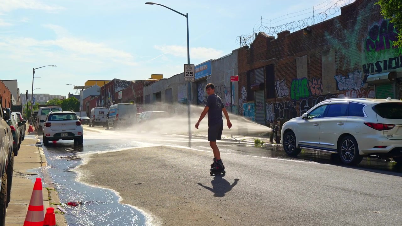 New York, USA, 1. juli 2026: Mand rulleskøjteløber gennem brandhanesprøjt på gaden. En rulleskøjteløber skøjter gennem vand sprøjtet fra en brandhane på en graffitibeklædt bygade i solskin