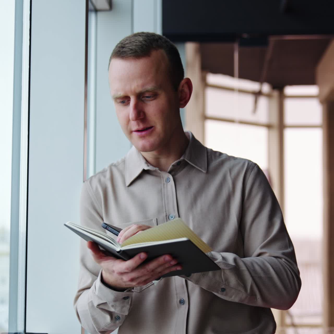 Young man leaned on the window holding a telephone and a paper notebook. Man turning pages in a book and comments something
