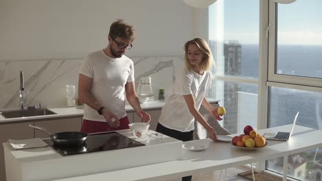 young attractive couple in domestic clothes on a bright studip kitchen - cooking simple breakfast.happy wife kissing her husband, man breaking eggs for an omelet. slow motion