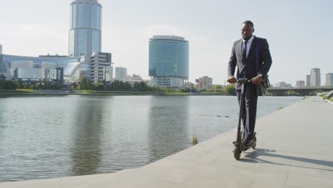 young serious businessman on electric scooter by riverside against tall modern buildings 1