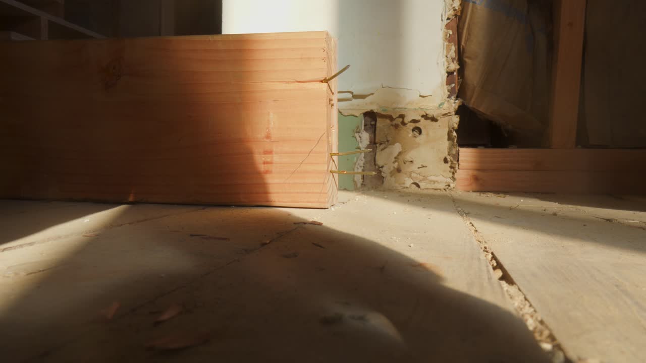 A close-up shot of a wooden beam lying on the floor in a construction environment. Nails protrude from the side of the beam.