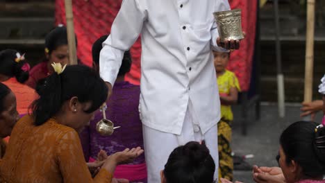 priest performs hindu ritual ceremony with water using ladle to pour into hands of followers seated on courtyard of the temple. women hold out hands to receive water blessing.