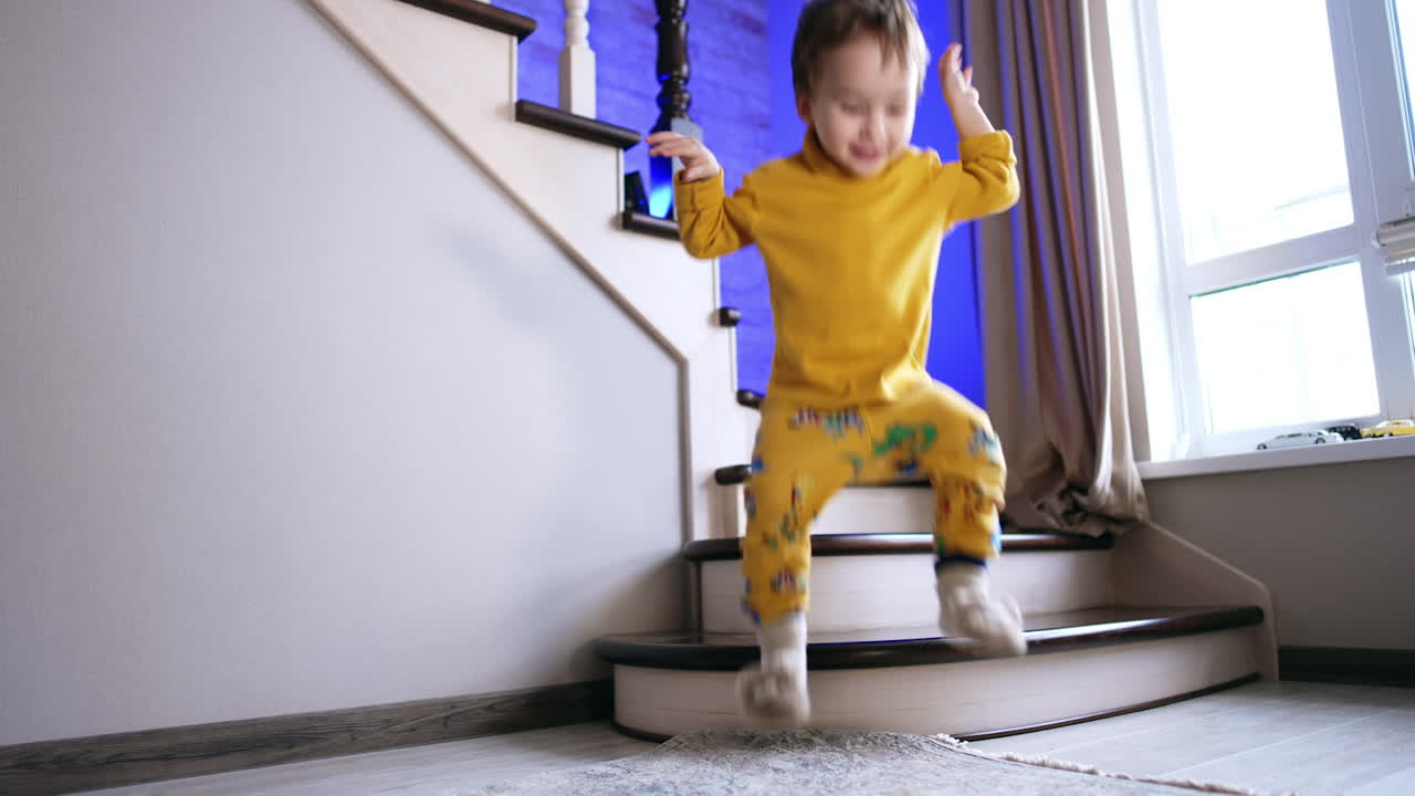 Two-year old toddler in yellow pajama stands on the stairs. Baby boy jumps cheerfully close to camera.